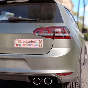 Op de afbeelding: Een roze bumpersticker met de tekst "I brake for critters" en illustraties van cartoon dieren. De sticker is rechthoekig en is bevestigd aan de achterkant van een zilveren auto.