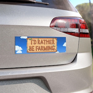 May include: A rectangular bumper sticker with the text "I'D RATHER BE FARMING." The design features a wooden sign with the text in brown letters, set against a blue sky with white clouds. The sticker is attached to the back of a car.