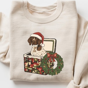 May include: Beige sweatshirt featuring a graphic of a dog wearing a Santa hat, sitting in a cooler. A Christmas wreath with a red bow is next to the cooler. The sweatshirt has long sleeves.