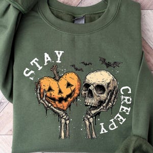 May include: A green sweatshirt with the words "STAY CREEPY" in white. The graphic features a jack-o'-lantern heart held by skeletal hands next to a skull, with bats above. The sweatshirt is folded on a wooden surface.