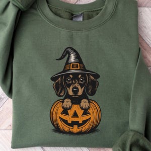 May include: Green sweatshirt featuring a dachshund wearing a witch hat and sitting inside an orange jack-o'-lantern. The graphic is in a cartoon style, perfect for Halloween. The sweatshirt has long sleeves and a crew neck.