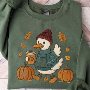 May include: Green sweatshirt featuring an embroidered duck wearing a hat and scarf, carrying a coffee cup, and walking past pumpkins. The design includes autumn leaves and an acorn. The sweatshirt has long sleeves.
