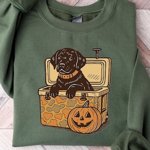 May include: Green sweatshirt featuring a black Labrador Retriever in a cooler with a pumpkin. The dog is wearing a collar. The cooler has a camouflage pattern. The pumpkin has a carved face. The sweatshirt has long sleeves.