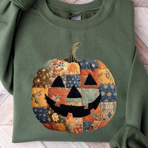 Peut inclure: Sweat-shirt vert olive avec un motif citrouille en patchwork. La citrouille est composée de divers motifs de tissu orange, bleu et jaune. Le sweat-shirt a un col rond et des manches longues, parfait pour l'automne.