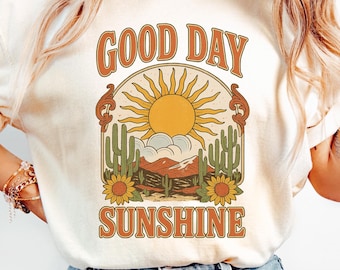 May include: Cream-colored t-shirt with the words "GOOD DAY SUNSHINE" in orange lettering. The graphic features a sun, cacti, and sunflowers. The model is wearing light blue jeans.