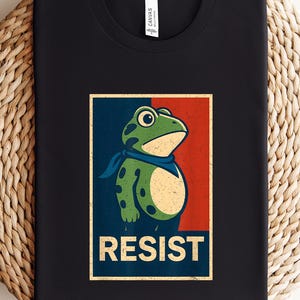 May include: Black t-shirt with a cartoon frog wearing a blue bandana, set against a red and blue background. The word "RESIST" is printed in a bold, vintage-style font below the frog illustration. The shirt is displayed on a woven surface.