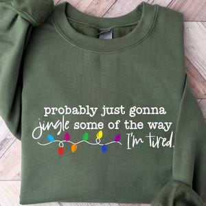 Puede incluir: Una sudadera verde oliva con el texto "probably just gonna jingle some of the way I'm tired" en escritura blanca. Debajo, una cadena de luces navide&ntilde;as de colores. La sudadera est&aacute; doblada sobre una superficie de madera clara.