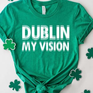 May include: A green t-shirt with the words "DUBLIN MY VISION" in white block letters. The shirt is surrounded by green shamrock decorations on a white background. The shirt is knotted at the bottom.