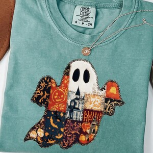 May include: A teal Comfort Colors t-shirt featuring a patchwork ghost design with black oval eyes and a white face. The ghost is made of various patterned fabrics with Halloween motifs. A silver necklace with a small pendant rests on the shirt.