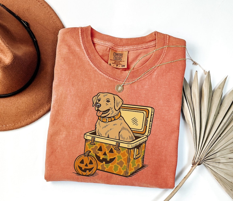 Puede incluir: Camiseta naranja Comfort Colors con un perro de dibujos animados en una nevera y una calabaza de Halloween. El perro lleva collar. La nevera tiene un estampado de camuflaje. Un sombrero marr&oacute;n y un collar tambi&eacute;n est&aacute;n en la imagen.