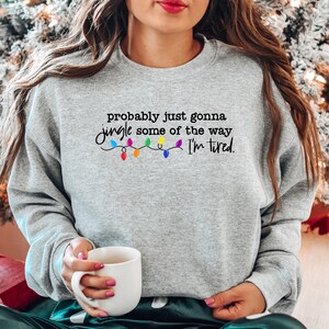 Puede incluir: Sudadera gris claro con la frase "probably just gonna jingle some of the way I'm tired." en escritura negra, con luces navide&ntilde;as coloridas. Una persona sostiene una taza blanca.