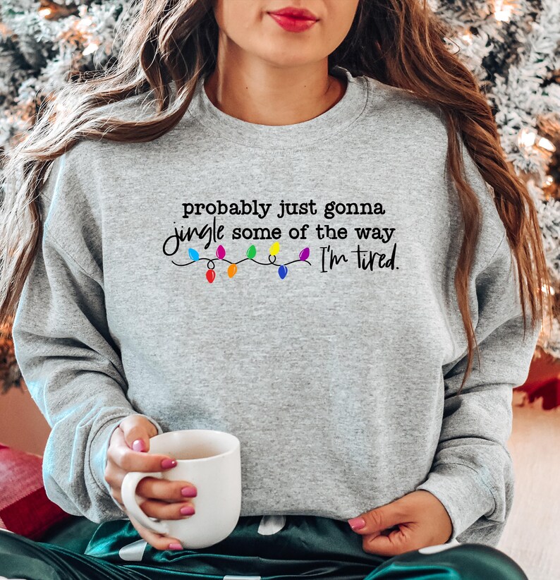 Puede incluir: Sudadera gris claro con la frase "probably just gonna jingle some of the way I'm tired." en escritura negra, con luces navide&ntilde;as coloridas. Una persona sostiene una taza blanca.