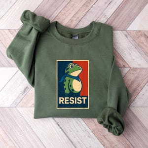 May include: An olive green sweatshirt with a graphic of a frog wearing a blue scarf, set against a red and blue background. The word "RESIST" is printed below the frog. The sweatshirt is folded on a wooden surface.