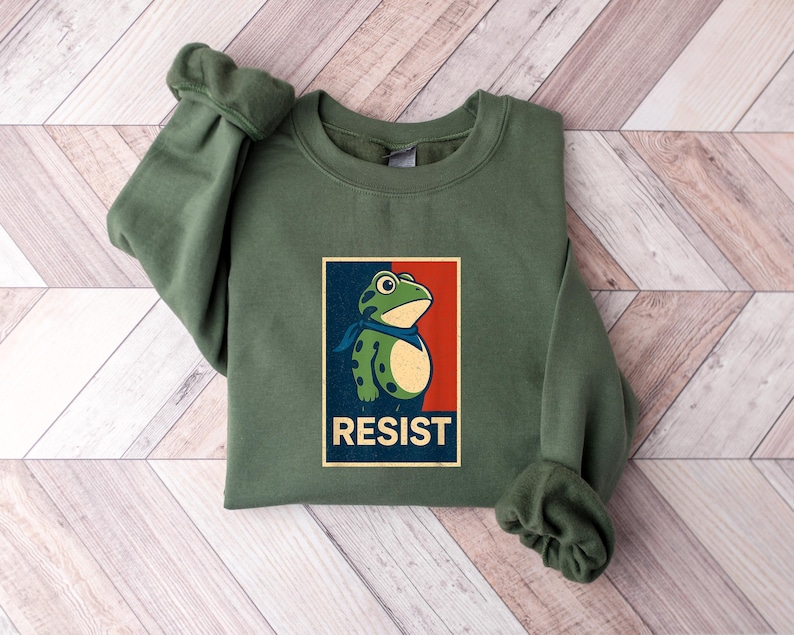 May include: An olive green sweatshirt with a graphic of a frog wearing a blue scarf, set against a red and blue background. The word "RESIST" is printed below the frog. The sweatshirt is folded on a wooden surface.