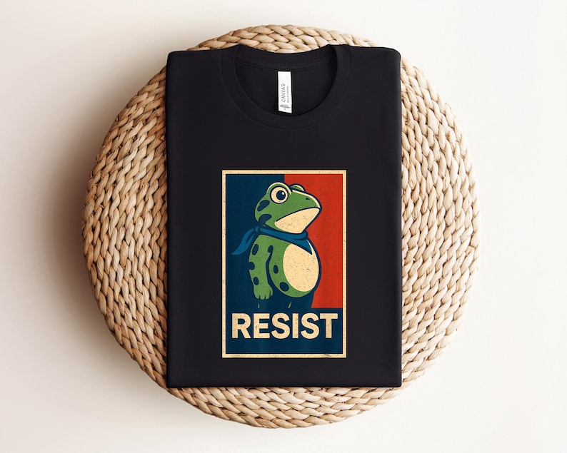 May include: Black t-shirt featuring a cartoon frog with a blue bandana, set against a red and blue background. The word "RESIST" is printed in a bold, vintage-style font below the frog illustration. The shirt is displayed on a woven surface.