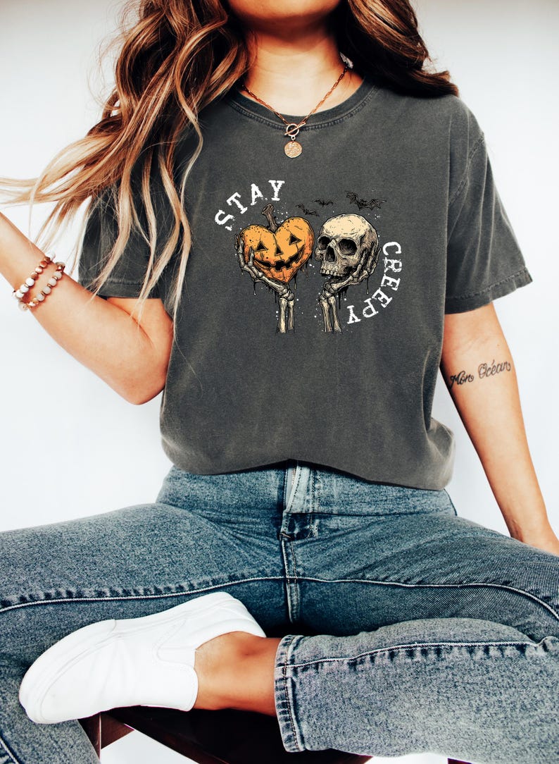 May include: Dark gray t-shirt with a Halloween-themed graphic. The design features a heart-shaped jack-o'-lantern held by skeletal hands next to a skull, with the words "STAY CREEPY" in a circular arrangement.