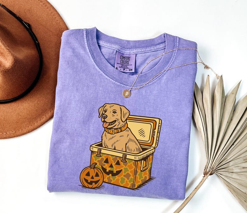 Puede incluir: Camiseta Comfort Colors de color lila claro con un perro de dibujos animados en una nevera y una calabaza. El perro es de color marr&oacute;n claro con un collar marr&oacute;n. La nevera tiene un patr&oacute;n de camuflaje y una cara de calabaza. Un collar dorado est&aacute; sobre la camisa.