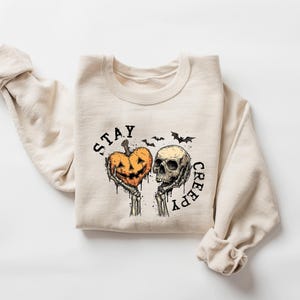 May include: Beige sweatshirt with a Halloween-themed graphic. The design features a jack-o'-lantern heart held by skeleton hands next to a skull, with the words "STAY CREEPY" and bats. A cozy, long-sleeved top.