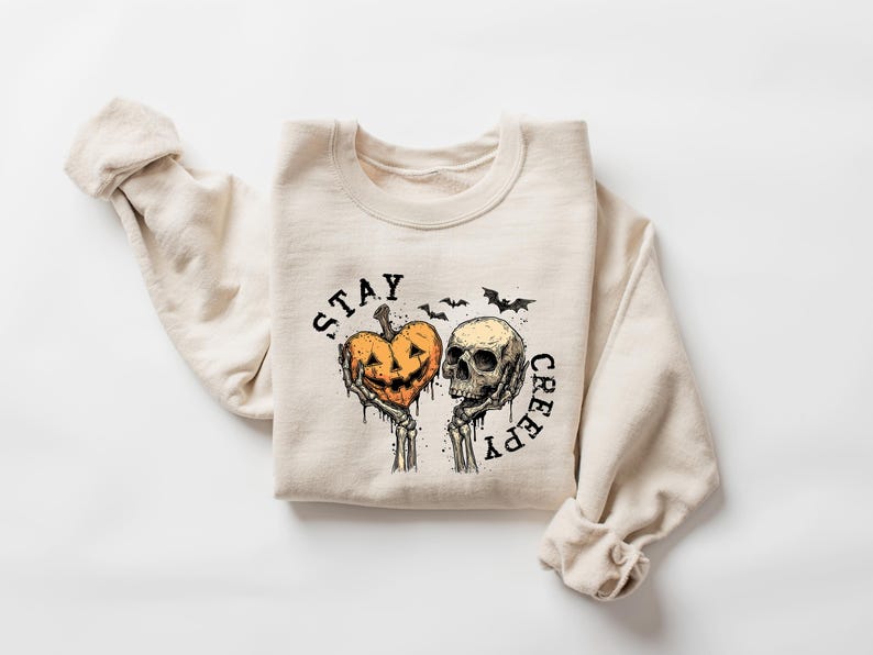 May include: Beige sweatshirt with a Halloween-themed graphic. The design features a jack-o'-lantern heart held by skeleton hands next to a skull, with the words "STAY CREEPY" and bats. A cozy, long-sleeved top.
