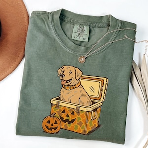 Puede incluir: Camiseta verde oliva con un perro Labrador Retriever de dibujos animados dentro de una nevera y una calabaza. La nevera tiene un patr&oacute;n de camuflaje. Un collar dorado est&aacute; drapeado sobre la camisa.
