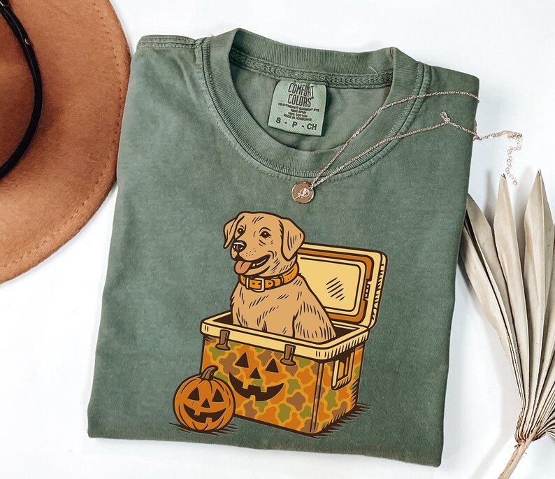 Puede incluir: Camiseta verde oliva con un perro Labrador Retriever de dibujos animados dentro de una nevera y una calabaza. La nevera tiene un patr&oacute;n de camuflaje. Un collar dorado est&aacute; drapeado sobre la camisa.