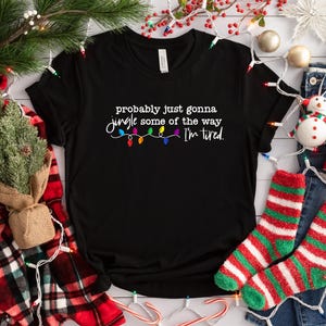 Puede incluir: Camiseta negra con el texto "probably just gonna jingle some of the way I'm tired" y una cadena de luces navide&ntilde;as de colores. La camiseta est&aacute; rodeada de adornos navide&ntilde;os, incluyendo calcetines, una taza y un peque&ntilde;o &aacute;rbol.