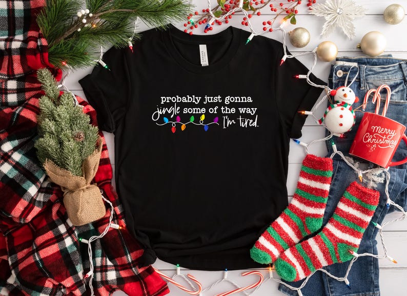 Puede incluir: Camiseta negra con el texto "probably just gonna jingle some of the way I'm tired" y una cadena de luces navide&ntilde;as de colores. La camiseta est&aacute; rodeada de adornos navide&ntilde;os, incluyendo calcetines, una taza y un peque&ntilde;o &aacute;rbol.