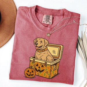 Puede incluir: Una camiseta Comfort Colors granate con un perro Labrador Retriever de dibujos animados dentro de una nevera y una calabaza. La nevera tiene un patr&oacute;n de camuflaje. Un collar plateado con un colgante est&aacute; en la camisa.