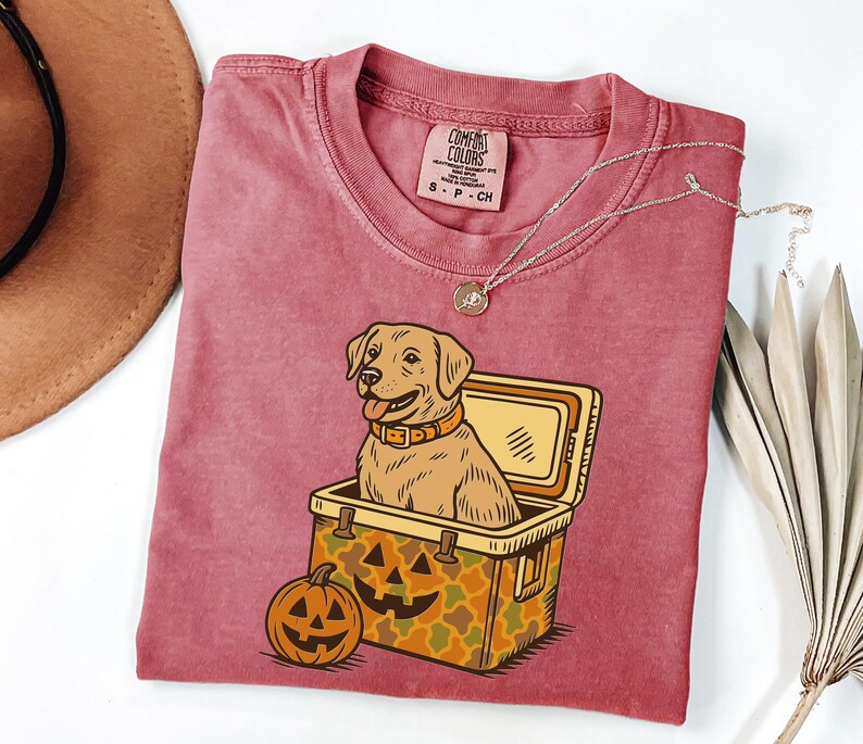 Puede incluir: Una camiseta Comfort Colors granate con un perro Labrador Retriever de dibujos animados dentro de una nevera y una calabaza. La nevera tiene un patr&oacute;n de camuflaje. Un collar plateado con un colgante est&aacute; en la camisa.