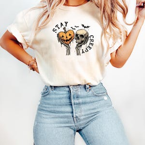 May include: Cream-colored t-shirt with a Halloween-themed graphic. The design features a jack-o'-lantern and a skull held by skeletal hands, with the words "STAY CREEPY" in a circular arrangement. Two bats are above the skull.