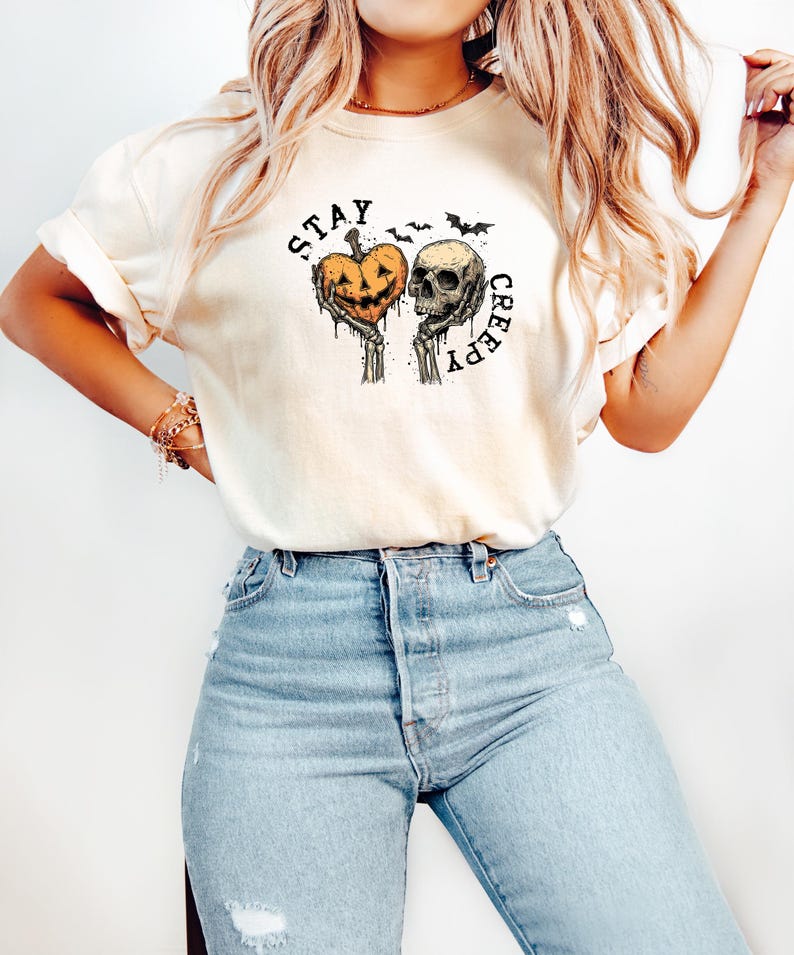May include: Cream-colored t-shirt with a Halloween-themed graphic. The design features a jack-o'-lantern and a skull held by skeletal hands, with the words "STAY CREEPY" in a circular arrangement. Two bats are above the skull.