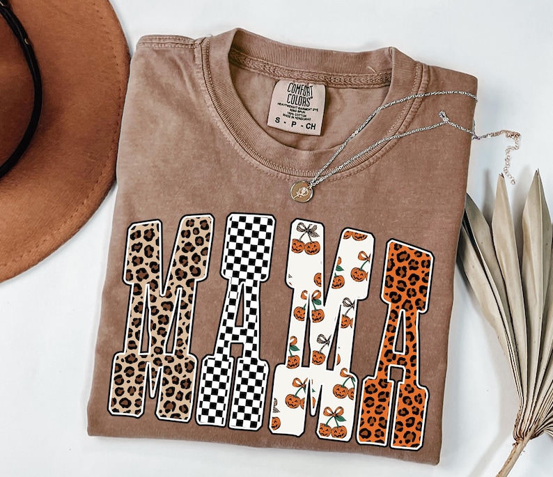 May include: A brown Comfort Colors t-shirt with the word "MAMA" in a variety of patterns, including leopard print, checkered, and pumpkin designs. A silver necklace with a small pendant is draped across the shirt.