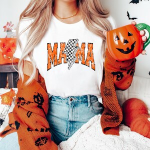 Pu&ograve; includere: T-shirt bianca con la scritta "MAMA" in lettere arancioni sbiadite e un fulmine a scacchi bianco e nero. La persona indossa un maglione arancione bruciato con disegni neri a tema Halloween e tiene in mano una tazza di ceramica a forma di zucca.