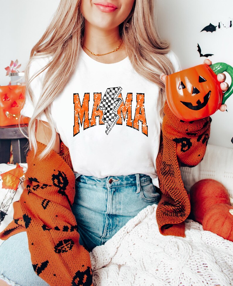 Pu&ograve; includere: T-shirt bianca con la scritta "MAMA" in lettere arancioni sbiadite e un fulmine a scacchi bianco e nero. La persona indossa un maglione arancione bruciato con disegni neri a tema Halloween e tiene in mano una tazza di ceramica a forma di zucca.