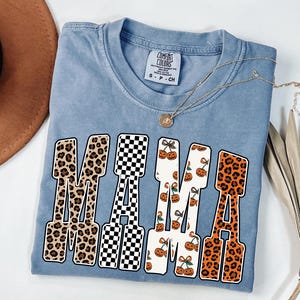 May include: A light blue Comfort Colors t-shirt with the word "MAMA" in a bold, block letter design. The letters feature leopard print, a black and white checkerboard pattern, and a pumpkin and cherry print. A gold necklace is draped across the shirt.