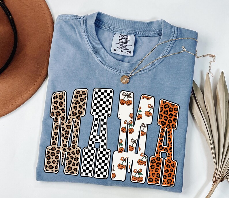 May include: A light blue Comfort Colors t-shirt with the word "MAMA" in a bold, block letter design. The letters feature leopard print, a black and white checkerboard pattern, and a pumpkin and cherry print. A gold necklace is draped across the shirt.