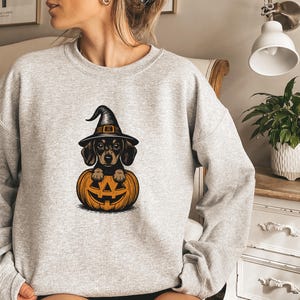 May include: A light grey sweatshirt featuring a Halloween-themed graphic. The graphic depicts a dachshund wearing a witch hat, peeking out from a carved orange pumpkin. The design is in a cartoon style.