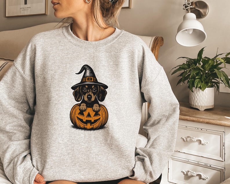 May include: A light grey sweatshirt featuring a Halloween-themed graphic. The graphic depicts a dachshund wearing a witch hat, peeking out from a carved orange pumpkin. The design is in a cartoon style.
