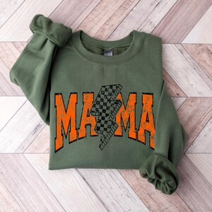 Pu&ograve; includere: Una felpa verde oliva con la scritta "MAMA" in lettere arancioni. Un fulmine a scacchi in bianco e nero &egrave; al centro della parola "MAMA". La felpa &egrave; piegata su una superficie di legno marrone chiaro.
