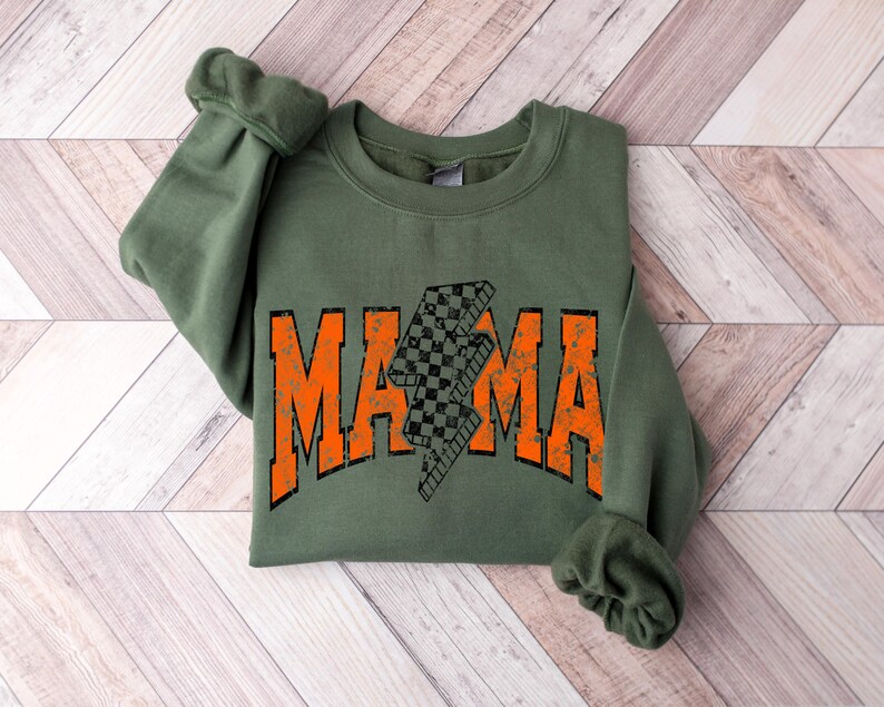 Pu&ograve; includere: Una felpa verde oliva con la scritta "MAMA" in lettere arancioni. Un fulmine a scacchi in bianco e nero &egrave; al centro della parola "MAMA". La felpa &egrave; piegata su una superficie di legno marrone chiaro.