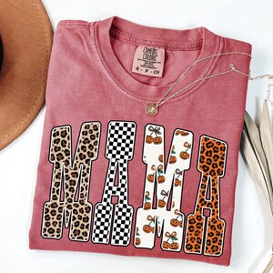 May include: A red-pink Comfort Colors t-shirt with the word "MAMA" in a bold, retro font. The letters feature leopard print, a black and white checkerboard pattern, and a cherry and pumpkin design. A gold necklace rests on the shirt.
