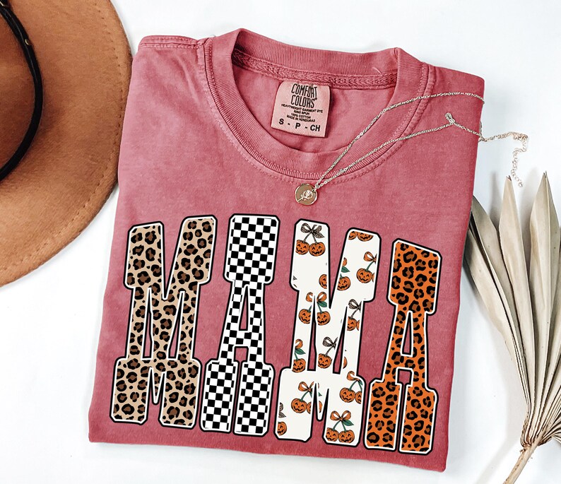 May include: A red-pink Comfort Colors t-shirt with the word "MAMA" in a bold, retro font. The letters feature leopard print, a black and white checkerboard pattern, and a cherry and pumpkin design. A gold necklace rests on the shirt.