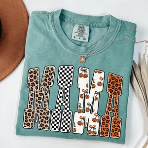 May include: A teal Comfort Colors t-shirt with the word "MAMA" in a bold, block letter design. The letters feature various patterns, including leopard print, black and white checkerboard, and cherry and pumpkin patterns. A silver necklace with a pendant is draped across the shirt.