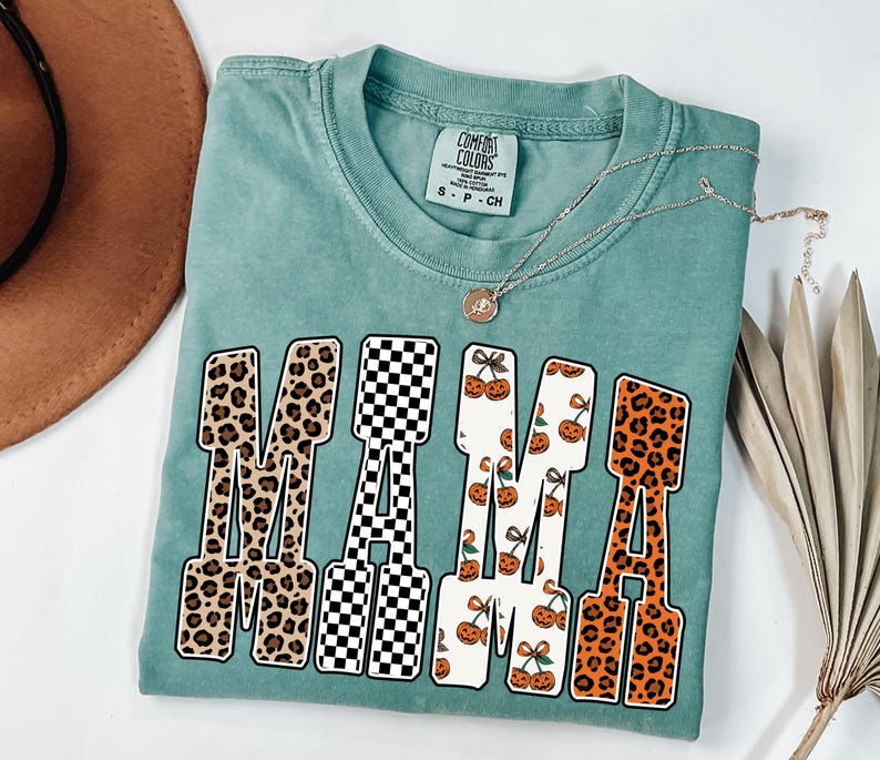 May include: A teal Comfort Colors t-shirt with the word "MAMA" in a bold, block letter design. The letters feature various patterns, including leopard print, black and white checkerboard, and cherry and pumpkin patterns. A silver necklace with a pendant is draped across the shirt.