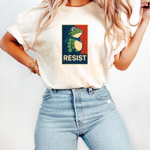 May include: Cream-colored t-shirt featuring a frog graphic with a blue scarf, set against a red and blue background. The word "RESIST" is printed below the frog. The shirt is paired with light blue jeans.