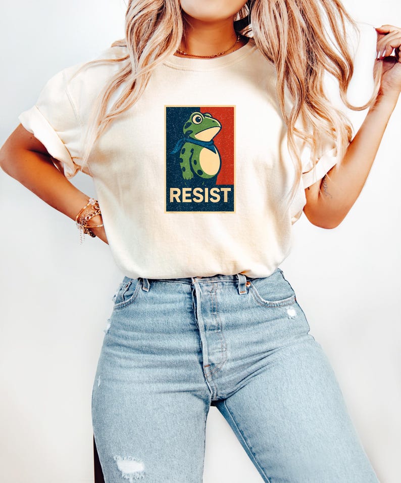 May include: Cream-colored t-shirt featuring a frog graphic with a blue scarf, set against a red and blue background. The word "RESIST" is printed below the frog. The shirt is paired with light blue jeans.