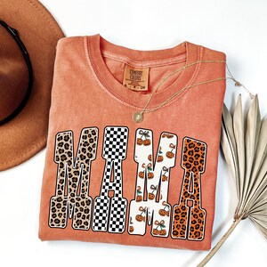May include: A coral-colored t-shirt with the word "MAMA" in a bold, patterned design. The letters feature leopard print, a black and white checkerboard, and a white background with orange pumpkin graphics. A gold necklace and a brown hat are also visible.