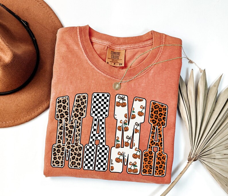 May include: A coral-colored t-shirt with the word "MAMA" in a bold, patterned design. The letters feature leopard print, a black and white checkerboard, and a white background with orange pumpkin graphics. A gold necklace and a brown hat are also visible.
