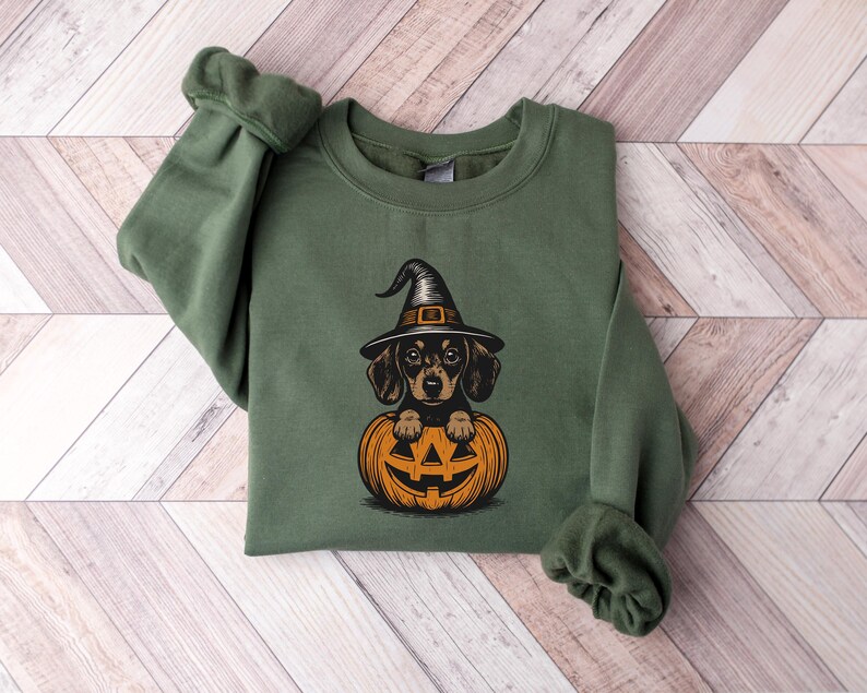 May include: Green sweatshirt featuring a dachshund wearing a witch hat and sitting inside an orange jack-o'-lantern. The graphic is in a cartoon style, perfect for Halloween. The sweatshirt has long sleeves and a crew neck.