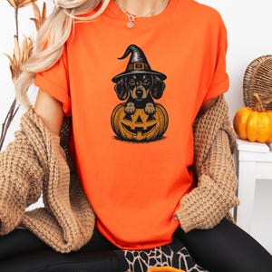 May include: Orange t-shirt featuring a black and tan dachshund wearing a witch hat, sitting inside a carved jack-o'-lantern. The design is a Halloween theme. The shirt is made of a soft material.